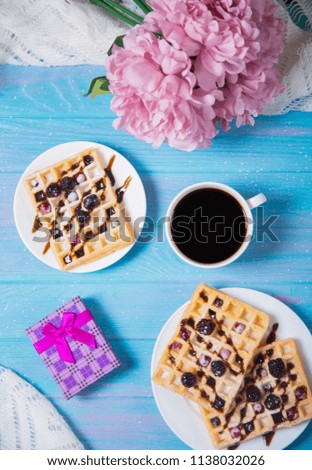 Morning composition with cup of coffee, wafers with berries, gift box and tender bouquet of beautiful pink peonies on blue wooden background. Concept of romantic breakfast, top view.