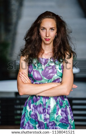 A head shot portrait of a young, attractive and beautiful Caucasian woman in the city against some grey steps. She is tall, slim and wearing a floral dress. She is smiling for the photo. 