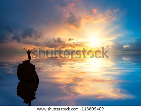 A man standing at lonely rock in the ocean with open arms set against a beautiful sunrise, clouds reflection in water