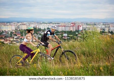 Two positive and energetic sportsmen riding bicycle on trail in high grass. Beautiful and cheerful man and woman in sportswear and helmets. Behind them city and built environment. Royalty-Free Stock Photo #1138185875