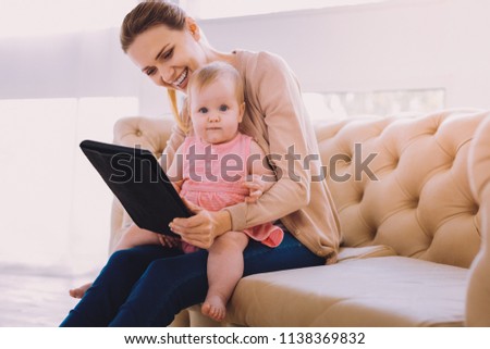 Progressive babysitter. Lovely baby girl sitting on the knees of a babysitter and looking at the pictures in her modern tablet