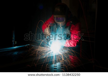 Metal worker in red working clothes welding steel in production factory on dark industrial background