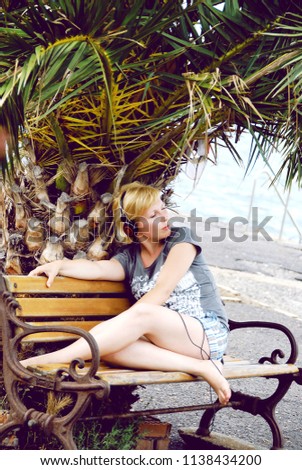blond girl listening to music on headphones under a palm tree on a bench. shore of the sea, brach, rest, summer. Royalty-Free Stock Photo #1138434200