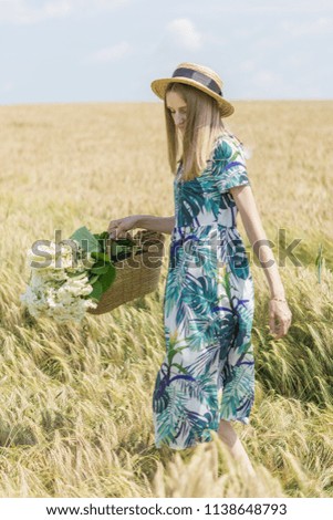 Beautiful young model in the field among the ears. The girl is blonde in a beautiful dress with a straw bag in which flowers