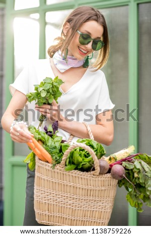 Portrait of a young woman with bag full of fresh raw vegetables standing outdoors near the green wall