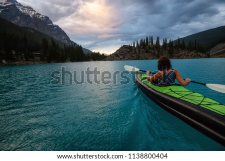 Adventurous woman kayaking in Moraine Lake during a striking cloudy sunset. Taken in Banff National Park, Alberta, Canada.