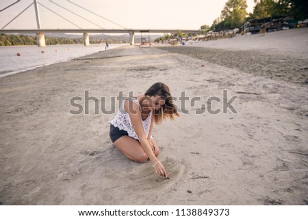 one young beautiful girl, drawing in sand with a stick, sitting in sand. Large beach in background with bridge. Novi Sad, Serbia.