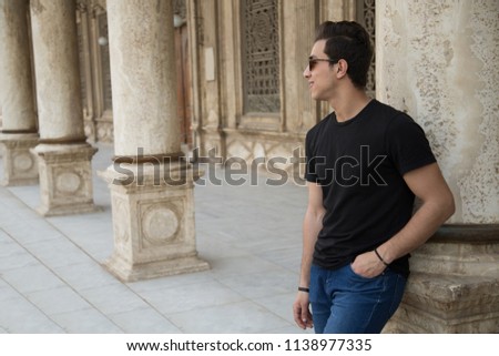 portrait of a young man wears a casual outfit and sunglasses, standing and leaning his back on column, looking away