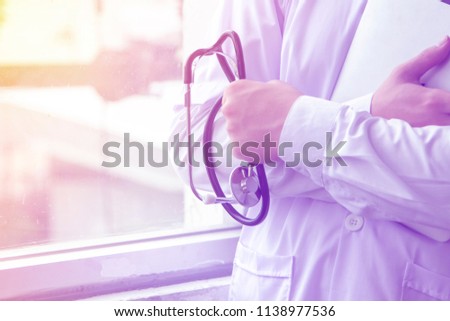 A doctor holds a stethoscope, medical instrument for examining heart beat,  and a file in front of a window thinking about his patient with orange and red light shine Royalty-Free Stock Photo #1138977536