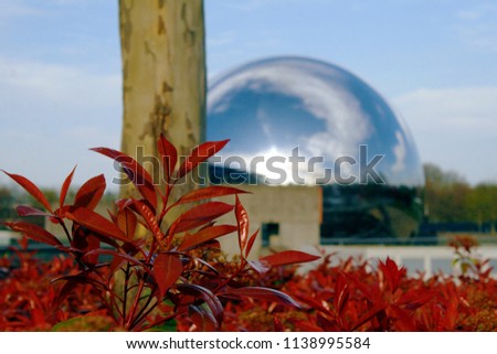 Giant metallic sphere with a mirror finish in the streets of Paris, composed in a picture with flowers and a tree from the garden surrounding that place.
