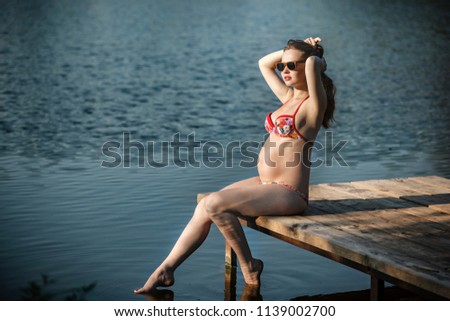 Attractive pregnant woman sits on the lake with sunglasses and a swimsuit.