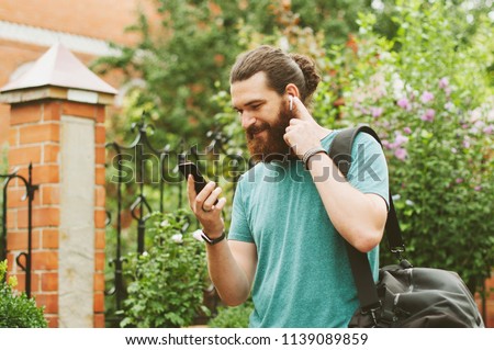 Cheerful happy bearded man looking at his smartphone, listening to the music with earpods outdoors. Royalty-Free Stock Photo #1139089859