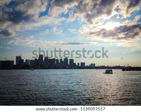 Distance view of Boston, Massachusetts, USA, harbor at the sun set from a boat. The sky is full of dark clouds and the sun evening light creates a strong contrast with the silhouette of the city.