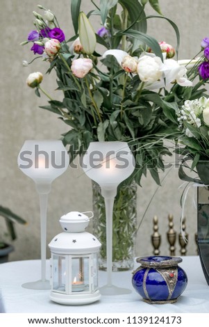 Interior composition with white glass candlesticks, white lantern, blue vintage utensil and peony, lily and lisianthus bouquet in background.