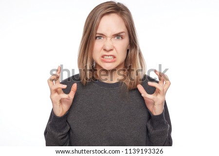 Light -haired annoyed angry woman holding hands in furious gesture. Young female in grey casual jumper expresses negative emotions, feelings, clenches teeth in anger, frowns face. Human being concept.