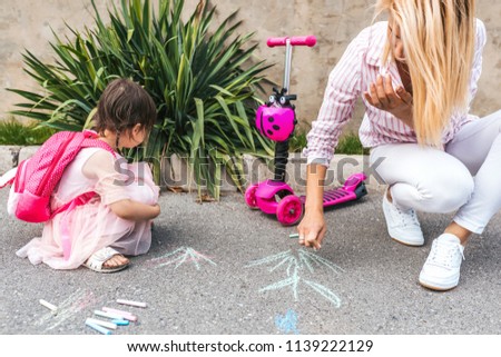 Image of happy little girl wears pink dress and mother drawing with colorful chalks on the sidewalk. Caucasian female play together with kid preschooler with backpack outdoor. Mom and child activity