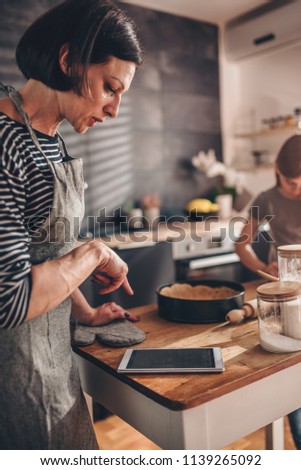 Mother and daughter standing in the kitchen by the wooden table kneading dough and searching apple pie recipe on the tablet