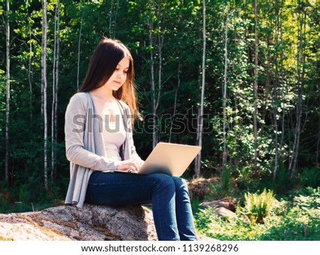 Young beautiful girl is working on a laptop while sitting on a big rock in the forest. Freelance concept