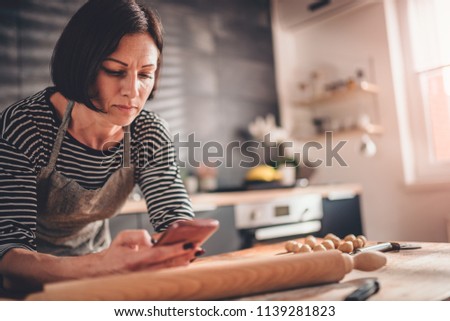 Woman standing in the kitchen by the wooden table and searching apple pie recipe on the smart phone