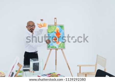 Teacher Man wearing black glasses holding a paintbrush is suggesting to drawing a goldfish.