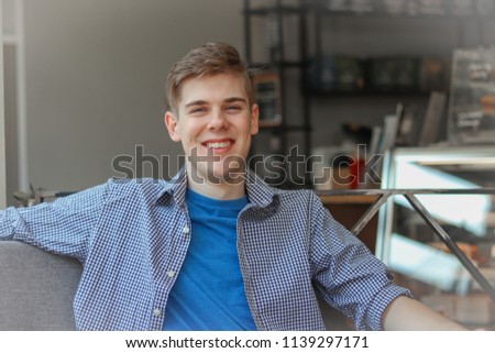 A portrait of a young man that is inside a building, smiling happy on a couch looking very casual. He is wearing a button down and blue t shirt.
