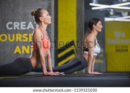 Focus on calm attractive fit lady bending back while staying on mat. She is stretching with her female friend together in gym. Her sporty mate is doing same exercise and smiling in background
