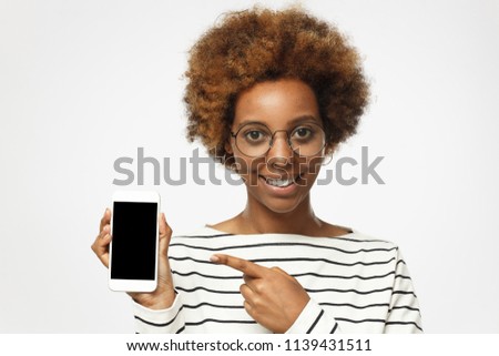Happy african american woman wearing roung glasses, isolated on grey background demonstrating blank smartphone and pointing to screen. Copyspace for advertising products and services.