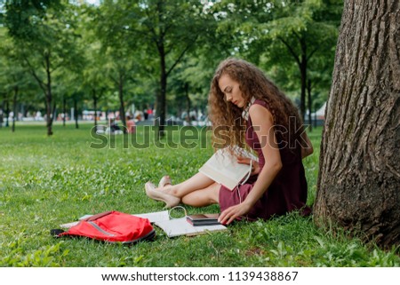 curly young beautiful girl student holding book and sitting under tree in summer park.