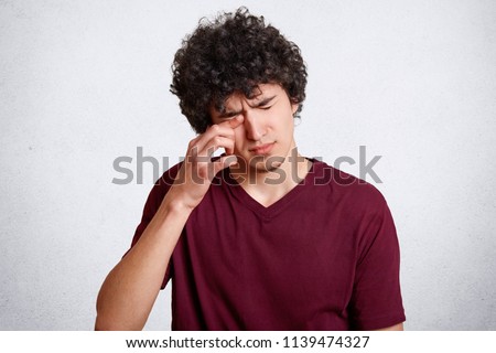 Tired teenager with crisp hair, rubs eyes as wants to sleep, has bad eyesight, dressed in red casual t shirt, isolated over white concrete wall. People, tiredness and health problems concept Royalty-Free Stock Photo #1139474327