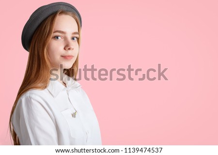 Adorable female with long straight hair, blue eyes, wears elegant black hat and white shirt, stands sideways, looks with intelligent expression at camera, isolated over pink wall with copy space