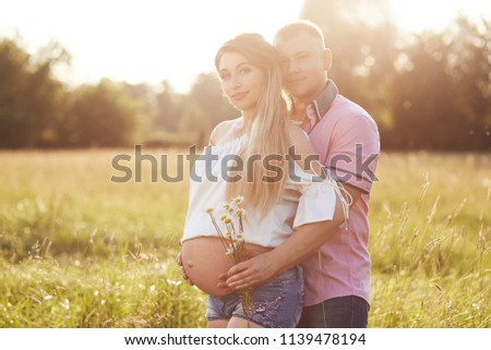 Attractive pregnant young female stands back to her affectionate husband, being happy future young parents, stand on field, enjoy sunny weather, love each other. Parenthood and pregnancy concept