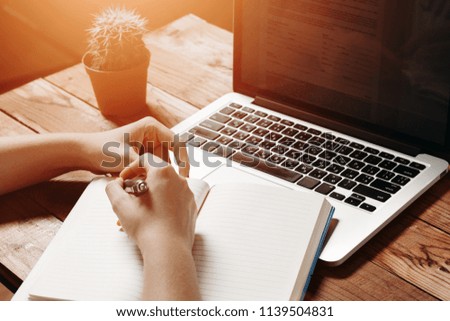 Female hands hold a notebook and write to it near laptop and flower on vintage wooden table. Mock-up with laptop and notebook 