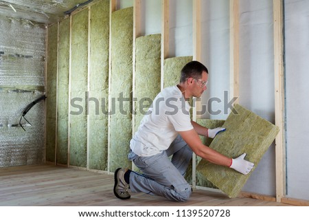 Worker in protective goggles and respirator insulating rock wool insulation in wooden frame for future house walls for cold barrier. Comfortable warm home, economy, construction and renovation concept Royalty-Free Stock Photo #1139520728