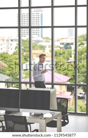 Young businessman holding a cup of coffee standing by the window. In a modern office There is a computer on the table, there is a bright green tree and the building is in the background.