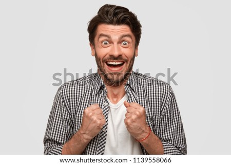 Wow, I done it finally! Glad positive young European male clenches fists with triumph, has happy look, anticipates for something pleasant, reacts on good news, isolated over white studio wall Royalty-Free Stock Photo #1139558066