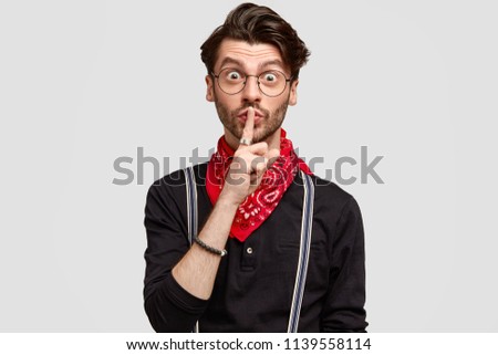 Surprised bearded male makes hush gesture, touches lips with index finger, looks secretly at camera, wears stylish black shirt with suspenders and red bandana, isolated over white background