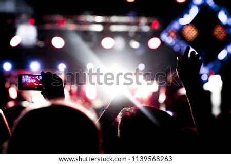 Audience with hands raised at a music festival and lights streaming down from above the stage. Soft focus, blurred movement