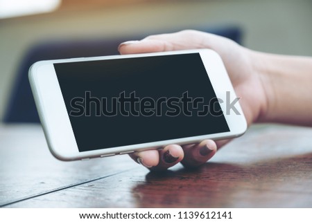 Mockup image of a woman holding and showing white mobile phone with blank black screen for watching on wooden table