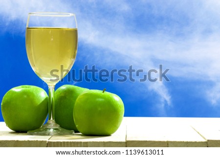 Green apple and glass of apple juice are on wooden floor front of blue sky background. 