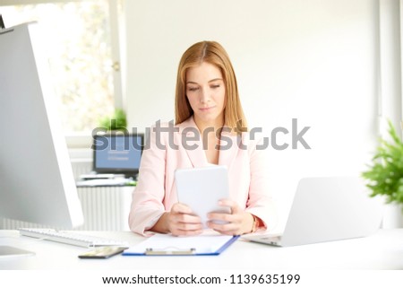 Attractive young businesswoman holding digital tablet in her hand and browsing on the internet while sitting at office desk and working.