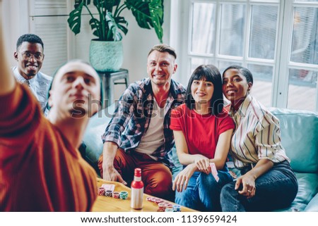 Happy multicultural hipster guys dressed in stylish casual wear sitting on comfortable couch and smiling while making photo during friendly meeting.Positive young people spending time together