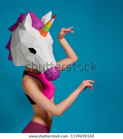 Young girl posing wearing unicorn's mask on saturated blue studio background. Horse's head mask made from paper having geometrical shape, bright colors. Slim body, pink panties, sport bra.