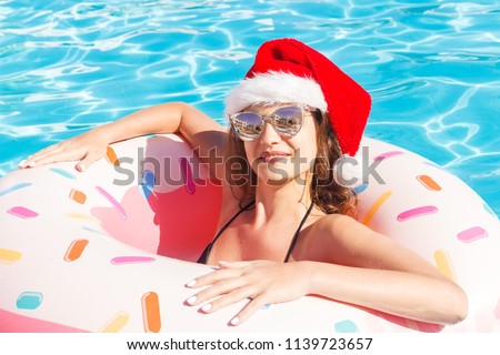 Beautiful young girl in bikini and Santa Claus hatwith donut inflatable pink circle in blue swimming pool.