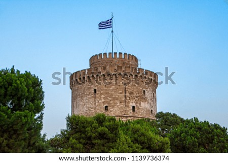 White Tower of Thessaloniki with Greek flag on top over the green conifer trees with blue sky background in Central Macedonia, Greece