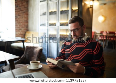 Restful man in casual sweater sitting by table in cafe, reading book and having drink at leisure
