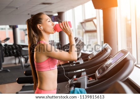 slim pretty joyful girl jogging at the treadmill and drinking a shaker with protein cocktail in the gym against the sunset. Concept of cardio exercises and healthy lifestyle Royalty-Free Stock Photo #1139815613