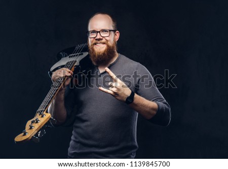 Happy redhead bearded male musician wearing glasses dressed in a gray t-shirt holds electric guitar and show rock and roll sign. Isolated on a dark textured background.