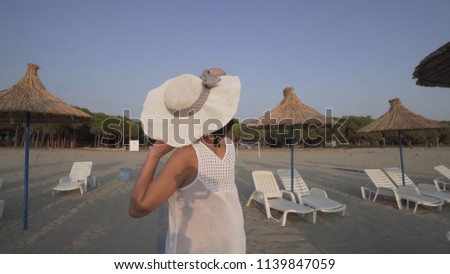 Happy smiling woman in white dress and hat blown by wind walking on empty beautiful beach with straw umbrellas and beds