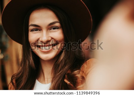Portrait of shiny girl taking picture. She is looking on camera and smiling. Girl wears brown hat. Also woman is holding camera with hand