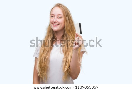 Blonde teenager woman holding credit card with a happy face standing and smiling with a confident smile showing teeth
